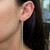 An image of a Messika brand earring with diamonds, worn on a woman's left ear. The earring is a vertical drop style featuring multiple small diamonds along its length, creating a line of shimmer down the side of her face. The photo is a close-up side view, showing the earring from the lobe to just above the shoulder, providing a clear look at the jewelry's design and how it dangles elegantly from the ear.