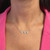 An image of a Messika diamond necklace for women, displayed on a model at a close frontal angle. The necklace features a delicate chain with evenly spaced diamonds and a centerpiece consisting of triangle-shaped diamond pendants, resting gracefully on the model's collarbone against a pink garment background.