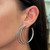 An image of a woman's ear showcasing a Messika earring with diamonds. The earring features a large hoop design with multiple diamond-encrusted bands, giving it a sparkling, layered appearance. The photo is taken in a close-up view with a side angle, focusing on the earring while it is being worn on the left ear. The background is blurred to emphasize the earring's details.