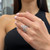 An image of a Messika brand ring with diamonds designed for women, showcased on a person's ring finger. The ring features a unique geometric design with multiple angles, and it is positioned approximately at the center of the image. The view is a close-up, focusing on the ring and the wearer's hand, which is placed against a blurred background, highlighting the intricate details and the sparkle of the diamonds. The person's hand is slightly angled towards the camera, allowing for a clear view of the top and side of the ring. The nails are painted a soft pink, complementing the white gold tone of the jewelry.