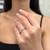 An image of a Messika women's diamond ring, delicately worn on the ring finger of a woman's left hand, which is positioned against a blurred background. The ring features a prominent, geometric design with sparkling diamonds set in a triangular pattern. The hand is slightly elevated and presented at a three-quarter angle to the camera, providing a clear view of the ring's intricate details from a medium distance.