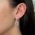 An image of a Messika diamond earring for women, showcased on a model's left ear. The earring features a close-up side view, exhibiting a dangling design with a geometric floral pattern, encrusted with multiple sparkling diamonds. The piece is angled to display both its intricate front design and its connection to the earlobe, with a medium distance allowing for clear visibility of the details and the earring's reflective quality.