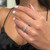 An image of a woman's hand with pale pink manicured nails, displaying a Messika brand ring with diamonds. The ring is worn on the ring finger, and the hand is positioned at a three-quarter angle to the camera, with a slight distance that allows the intricate details of the ring to be in focus against the soft, blurred background.
