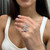 An image of a Messika women's ring with diamonds, worn on the ring finger of a person's left hand. The ring features an intricate design and is displayed prominently in the center of the image. The hand is positioned at a three-quarter angle, with the fingers slightly curled to show the ring's profile. The background is blurred, and the focus is on the ring, highlighting its details and the diamonds' sparkle. The distance between the camera and the ring is close enough to clearly capture the ring's elegance and craftsmanship.