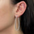 An image of a Messika brand earring for women, featuring a teardrop-shaped hoop design encrusted with diamonds. The earring is worn on the left ear, shown in a side view with the model facing to the right, providing a clear view of the earring's shape and the sparkling stones. The earring hangs just below the earlobe, and the photograph is taken at a close distance, focusing on the earring against the backdrop of the model's ear and hair.