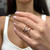 An image of a woman's hand displaying a Messika brand ring with diamonds. The ring is worn on the ring finger and features a prominent circular diamond setting surrounded by smaller diamonds. The hand is positioned against a blurred background with the fingers slightly bent, giving a clear view of the ring's details. The image is taken from a close, slightly elevated angle focusing on the top of the hand to emphasize the jewelry.