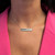 An image of a Messika necklace with diamonds worn by a woman, centered and close-up on her neck. The necklace features a horizontal bar pendant adorned with a row of sparkling diamonds, prominently displayed against a pink backdrop. The chain is thin and delicate, and the pendant sits just below the woman's collarbones, offering a clear view of the jewelry's design and brilliance.