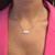 An image of a Messika diamond necklace for women, featuring a rectangular diamond pendant centered on a delicate chain. The necklace is positioned against the skin, resting just below the collarbone of a person wearing a pink top. The viewpoint is a close-up, frontal shot, highlighting the sparkle of the diamonds and the elegance of the piece.