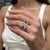 An image of a Messika women's ring with diamonds, showcased on a woman's ring finger. The ring features a unique geometric design with multiple diamond-encrusted bands intersecting at various angles. The photo is taken from a close, slightly elevated angle, emphasizing the sparkle of the diamonds and the intricate details of the ring. The background is softly blurred, drawing focus to the ring and the well-manicured hand wearing it.