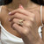 An image of a Messika women's ring with diamonds, showcased on a person's finger. The ring features a unique heart-shaped design with pavé diamonds, and is positioned roughly at the center of the frame at a close-up angle, highlighting its intricate details and craftsmanship. The background is softly blurred, focusing attention on the ring. The person is wearing a white sleeveless top, and only part of their hand and torso are visible, maintaining the emphasis on the sparkling ring.