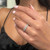 An image of a Messika diamond ring for women, showcased on a finger at a close angle, with the ring positioned at a slight tilt to display its circular diamond settings and intricate design details. The background is blurred, emphasizing the ring's sparkle against the wearer's manicured nails with light pink polish.