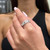 An image of a Messika brand women's ring with diamonds, prominently displayed on the ring finger of a hand with light pink polished nails. The hand is angled slightly towards the camera, giving a clear view of the ring's design and stones. The ring is in focus, set against a softly blurred background, emphasizing the sparkle of the diamonds. The distance from the camera allows for a detailed look at the ring's features while also capturing part of the hand and a hint of the black attire of the wearer.