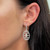 An image of a Messika earring with diamonds, designed for women, as worn on the left ear. The earring is in clear focus and is seen from a side angle, dangling just below the earlobe. It features a sparkling diamond-encrusted pendant with a geometric floral pattern, attached to a slender, diamond-set bar. The photo is a close-up, showcasing the intricate design and brilliance of the diamonds against the backdrop of the wearer's dark hair and skin tone.