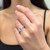 An image of a woman's hand gracefully positioned with the fingers slightly apart, showcasing a Messika ring with diamonds worn on the ring finger. The ring features a distinctive geometric design with sparkling diamonds set in a triangular pattern that catches the light. The close-up view emphasizes the ring's intricate details and the reflective quality of the stones. The hand is centered in the frame with soft focus on the background, drawing attention to the luxurious accessory.