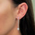 An image of a Messika brand Pend Glam'Azone drop earring for women, featuring diamonds. The earring is depicted in a close-up side view, worn on the left earlobe. It has a vertical orientation with a linear drop design, leading down to a larger round diamond at the bottom. The photo is taken at a slight angle, focusing on the earring from a medium distance, capturing the sparkle of the diamonds against the skin.