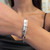 An image of a Messika brand Kate Sur chaine bracelet with diamonds, designed for women, worn on a wrist. The bracelet is positioned at the center of the frame, shown from a side angle, emphasizing its shine and the detail of the diamonds. The shot is taken at a close distance, providing a clear view of the bracelet's design and the sparkling diamonds on the upper half, while the lower half is attached to a delicate chain, both reflecting light on the wearer's skin.