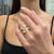 An image of a woman's hand displaying a Messika brand ring with diamonds. The ring features a unique geometric design with multiple triangular facets encrusted with sparkling diamonds. The hand is positioned in front of the wearer's body, with the ring centrally located on the ring finger. The image is taken from a close-up, frontal angle, emphasizing the intricate details and luxurious appearance of the ring. The background is softly out of focus to draw attention to the ring.
