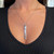 An image of a Messika Sautoir Kate Diamond Bar pendant necklace, designed for women. The necklace features a vertical bar pendant adorned with sparkling diamonds, centrally positioned against a black V-neck garment. The pendant hangs from a delicate white gold chain that rests at collarbone level. The shot is a close-up, taken from a front angle, highlighting the shine and details of the diamonds on the pendant.