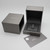 An image of an open Messika ring box positioned at the center, viewed from a top-down angle. There is a matching outer box partially visible to the left with the brand name "MESSIKA" prominently displayed. In front of the ring box lies a gray Messika card angled slightly to the right. The ring is not visible in the image, suggesting it may be absent or out of view due to the angle and distance of the shot. The background is a neutral white, emphasizing the dark tones of the box and card.