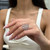 An image of a woman's hand displaying a Messika-brand Promess ring with diamonds. The hand is positioned slightly off-center to the left with fingers extended towards the right side of the frame, showcasing the ring at a close but comfortable distance. The ring features an intricate design with multiple bands and shines brightly, indicating high-quality diamonds and craftsmanship. The backdrop is softly focused, emphasizing the ring's detail.
