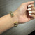 An image of a gold Rachel Koen bracelet with diamonds, worn on a woman's wrist. The bracelet features a design with interlocking folds and is positioned on the forearm approximately two inches from the wrist joint. The view is a close-up at a slight angle, emphasizing the bracelet's design and the diamonds' sparkle. The woman's hand is resting against a grey textured surface, with her fingers slightly curled inward and nails painted white, enhancing the bracelet's elegance.