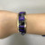 An image of a Black Jade Fire Opal Raoul Sosa women's bracelet with diamonds on a model's hand showing the proper positioning on the wrist. The bracelet features a 14k yellow gold band with inlaid black jade gemstones and opal fragments separated by thin yellow gold lines, accented with small, sparkling diamonds along one edge. The background is grey, highlighting the jewelry's colors and shine.