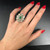 An image of a Vintage woman's hand with red polished nails showcasing a Rachel Koen ring with diamonds and emerald. The ring, worn on the ring finger, features a central green emerald surrounded by multiple sparkling diamonds set in a floral design. The hand is positioned against a dark background, with the ring displayed prominently at a close, slightly angled view that highlights the intricate details and the shine of the emerald.The condition of the ring is pre owned. 