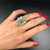 An image of a Vintage woman's hand wearing a Rachel Koen ring with diamonds and emerald. The ring features a prominent green emerald at the center surrounded by intricate designs and smaller diamonds. The hand is positioned against a dark background, with the fingers slightly spread to showcase the ring. The photo is taken from a close, top-down angle to give a clear view of the ring's details. The fingernails are painted red, contrasting with the silver tone of the ring.The condition of the ring is pre owned. 