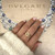 An image of a woman's hand adorned with a Bvlgari ring featuring diamonds, positioned at the center with a slight tilt towards the camera. The ring is on the ring finger, and the hand is resting against a backdrop with the Bvlgari logo and part of a decorative necklace in the background. The focus is on the ring, which is in the foreground, creating a sense of depth with the background slightly blurred.The condition of the ring is new. 