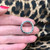 An image of a Bvlgari  B.Zero1 unisex adult ring without stones, held between two fingers at a slight angle. The ring is centered and close-up, showing engraved brand lettering. The background is out of focus with a leopard print pattern.The condition of the ring is pre-owned. 