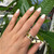 An image of a Bvlgari B.Zero1 ring with diamonds, worn on the ring finger of an adult's left hand, which is shown against a backdrop of green leaves. The hand is positioned with the palm facing inward and the fingers slightly separated, providing a clear view of the ring at a perpendicular angle to the camera. The ring features a multi-band design with diamond embellishments, capturing the light at a close, yet comfortable distance for detailed viewing.The condition of the ring is new. 