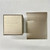 An image of a Bvlgari branded ring box and packaging, presented on a textured surface. The ring box, positioned on the left, is square, with a beige-gold finish and the Bvlgari logo embossed in the center. To the right, there is a rectangular packaging piece, standing upright with a matching beige-gold hue and the Bvlgari logo at the top. The items are captured from a top-down angle, with the camera at a close distance, providing a clear view of the textures and branding. The actual ring with diamonds is not visible in this image.