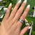 An image of a unisex adult's hand displaying a Bvlgari B.Zero1 ring with diamonds. The ring is worn on the ring finger and is positioned at a slight angle to the camera, offering a clear view of the band and the embedded diamonds. The hand is shown in close-up with a backdrop of green leaves, emphasizing the ring's design and sparkle. The condition of the ring is new. 