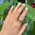 An image of a unisex adult's hand wearing a Bvlgari B.Zero1 ring with diamonds. The hand is positioned against a green leafy background, with the ring displayed prominently on the ring finger. The ring features multiple bands, some with diamond embellishments, viewed from a top-down angle at a close distance, giving a clear view of the ring's design and the sparkling diamonds.The condition of the ring is new. 