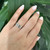 An image of a unisex Bvlgari B.Zero1 Openwork Logo Spiral one band ring without stones, shown on a person's ring finger, which is centered and in focus against a blurred background of green foliage. The ring is white gold with brand's signature design elements, and the hand is positioned at a slight angle, allowing a clear view of the top and side of the ring. The photo is taken from a close, but comfortable distance to highlight the detail of the ring.The condition of the ring is new. 