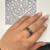 An image of a Bvlgari B.Zero1 unisex adult two band matt black ring without stones, shown worn on the ring finger of a hand with manicured nails. The hand is resting on a surface with a printed pattern resembling a diamond-encrusted watch face. The ring features a multi-band design with contrasting darker and lighter metallic tones. The perspective is a close-up, top-down view, focusing on the ring at a slight angle to capture the design details and the fit on the finger.The condition of the ring is new. 