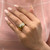 An image of a Rachel Koen brand women's ring with diamonds, showcased on a person's ring finger. The ring is positioned at a slight angle, with the diamonds catching the light prominently, and appears to be a close-up shot, emphasizing the ring's details against the backdrop of a pink fabric surface. The wearer's hand is elegantly posed with well-manicured, light pink nails, complementing the ring's luxurious appearance.