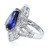 An image of a Rachel Koen women's ring with diamonds, showcasing a large, oval-cut blue Tanzanite set at an angle, flanked by intricate bands of smaller diamonds that sparkle against the silver setting. The ring is photographed from a side perspective, with the blue tanzanite prominently displayed in the foreground, occupying the upper left portion of the image, while the diamond-encrusted bands curve around it, leading to the polished white gold band that is visible in the background on the right side. The image is taken from a close distance, allowing for detailed observation of the ring's design and the gemstone's facets.