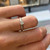 An image of a Tiffany & Co. engagement ring with diamonds, displayed on a woman's finger. The ring features a prominent square-cut diamond set in a platinum band. The photo is taken from a close, slightly elevated angle focusing on the top of the ring, with the background softly blurred to emphasize the ring's details.