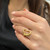 An image of a Tiffany & Co. women's ring without stones, featuring a heart-shaped design in yellow gold, showcased on a person's finger. The ring is positioned at a slight angle towards the camera, giving a clear view of the heart shape and the band. The photo is taken from a close distance, emphasizing the ring against the blurred background of the person's attire and the interior setting.