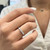 An image of a Rachel Koen brand women's ring with diamonds, showcased on a person's ring finger. The photo is a close-up shot with a slight top-down angle, emphasizing the sparkle of the diamonds which are set in a row on the band. The background is softly blurred, drawing focus to the ring, and the distance is close enough to detail the ring's design while showing the fingers and part of the person's white shirt.The ring is pre owned in a great condition.
