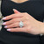 An image of a Rachel Koen women's ring with diamonds, prominently displayed on a woman's ring finger. The ring is viewed from a top-down angle at close distance, showcasing a large central diamonds with a halo of smaller diamonds and additional diamonds encrusting the band. The woman's hand is resting against a black fabric background, with her nails painted in a soft pink shade.The ring is pre owned in a great condition.