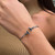 An image of a sleek Tiffany & Co. unisex adult bracelet, displayed on a person's wrist in white gold. The bracelet features a simple, elegant design with a polished finish and distinctive geometric details. The photo is taken at a close distance, with the bracelet in clear focus, positioned horizontally across the wrist. The angle of the shot provides a clear view of the bracelet's design and how it sits on the skin, against a blurred background of the person's neutral-colored clothing and bare shoulder.