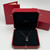 An image of a Cartier Double C De Cartier women's necklace with diamonds, displayed in an open red Cartier box with a black velvet interior. The necklace features a delicate rose gold chain with a diamond-studded pendant, centered and hanging at a straight angle. The box is positioned at a slight left-facing angle and is in the foreground, with two Cartier boxes blurred in the background. The viewpoint is from a moderate distance, capturing the entire open box and a portion of the background boxes. Comes in a great preowned condition.