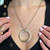 An image of a Cartier Tri Color Trinity Panthere Pendant necklace with diamonds displayed on a woman's neck. The necklace features a large, circular gold pendant with a concentric design, encrusted with diamonds along its circumference. The pendant hangs centrally and is attached to a fine gold, rose and white gold chain. The view is a close-up at a straight-on angle, focusing on the necklace against the backdrop of a black garment, with the woman's hands gently touching the pendant, showcasing pale pink manicured nails. The photo is taken from a short distance, capturing the details of the necklace and the upper chest area. Comes in a great preowned condition.