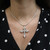 An image of a Rachel Koen diamond necklace for women, featuring a prominent cross pendant with diamond embellishments. The necklace is displayed on a person, centered and hanging at a short drop length, with the cross positioned just above the sternum. The chain enters the frame from the top corners, suggesting a close-up frontal view. The background shows the person wearing a black garment with white polka dots, adding contrast to highlight the necklace.