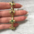 An image of a gold Catherine Mullen women's bracelet with diamonds and gemstones, displayed vertically across a person's fingers to provide a clear view. The bracelet features alternating gemstone settings with a mix of sapphires, emeralds, and ruby, each separated by diamond-encrusted links. The angle is a close-up, focusing on the bracelet, which is held at a short distance from the camera, with a marble surface in the background.