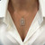 An image of a necklace with diamonds and gemstones by Rachel Koen, designed for women. The necklace features a rectangular pendant with intricate detailing, centered against a white blouse. The image is taken from a front angle, focusing closely on the jewelry to highlight its elegance and craftsmanship.Excellent pre-owned condition. 