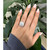 An image of a woman's hand wearing a Bottega Veneta ring with diamonds. The hand is positioned palm down, with fingers slightly spread, showcasing the ring on the ring finger. The angle is slightly tilted from above, capturing the ring's intricate diamond design. The distance is close-up, focusing on the hand against a backdrop of large green leaves.