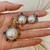 An image of a women's jewelry set by Stephen Dweck, featuring a pendant and matching earrings. The set is held in a hand, positioned horizontally. The pendant and earrings are adorned with large central pearls, surrounded by smaller, round, purple amethyst set in yellow gold. The photo is taken from a close angle, providing a clear view of the intricate design and details.Its excellent pre-owned condition. 